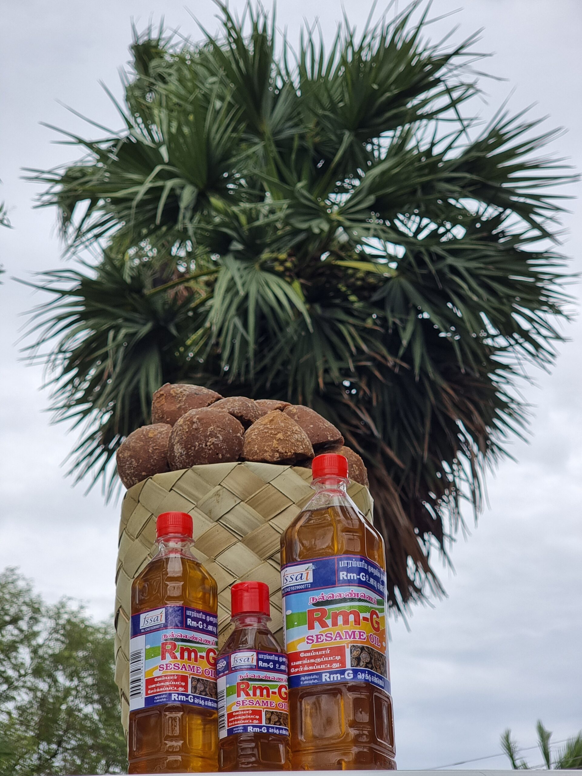 Unrefined Sesame Chekku Oil Bottles of Rm-G Sesame Unrefined Chekku Oil are placed beside a basket of palm jaggery, with a palm tree in the background under a cloudy sky.